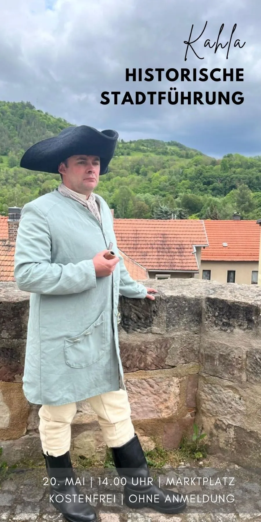 Mann in historischer Kleidung mit Dreispitz und Pfeife lehnt an einer Steinmauer vor grünen Hügeln und Häuserdächern.